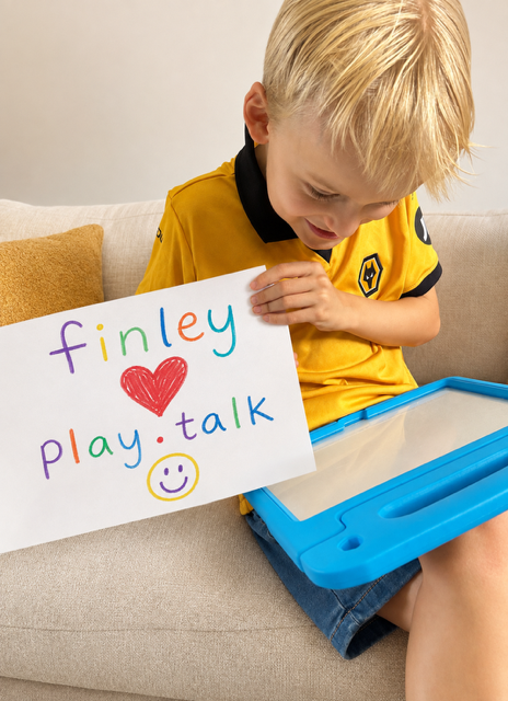 Finley on the sofa with his iPad, holding up a hand-drawn sign that reads finley, a heart, play.talk